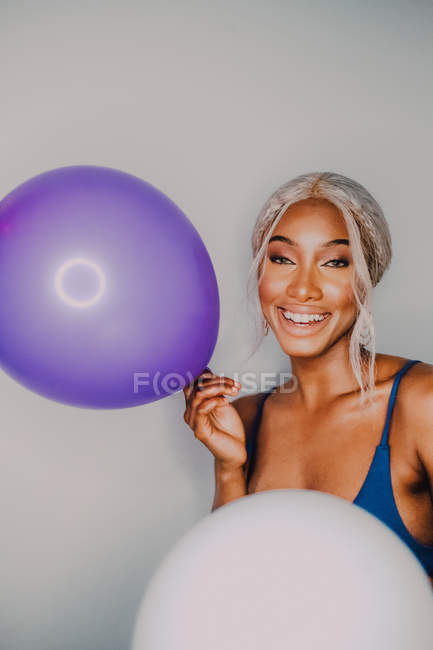 Gioiosa donna adulta nera con palloncini colorati guardando la fotocamera mentre in piedi su sfondo bianco — Foto stock