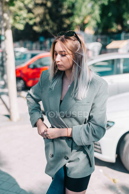 Businesswoman with trendy hairstyle and suit holding laptop with legs and looking away in parking lot at bright day — Stock Photo