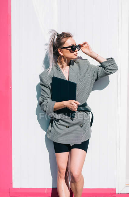 Empresária com penteado na moda, óculos de sol e terno segurando laptop e olhando para longe na parede — Fotografia de Stock