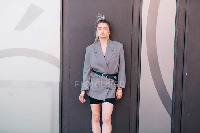 Trendige Frau mit Modefrisur und Kostüm, die in die Kamera blickt, die an die Tür gelehnt ist — Stockfoto