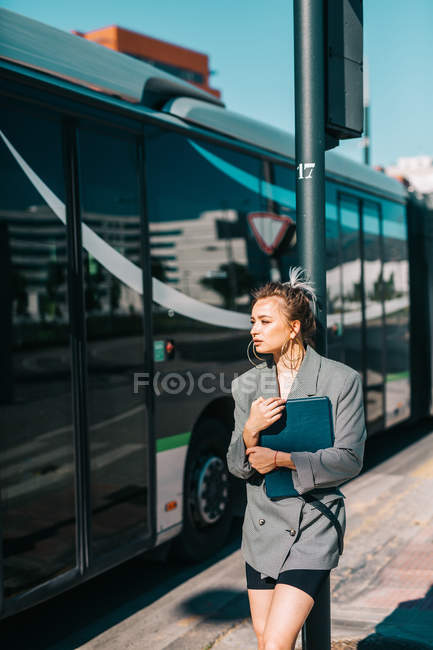 Donna d'affari alla moda che tiene tablet con vestito e occhiali da sole appoggiati alla fermata dell'autobus e guardando altrove — Foto stock