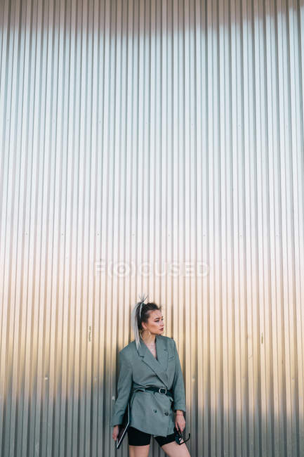 Donna d'affari alla moda che tiene tablet e occhiali da sole, appoggiata alla parete metallica e distogliendo lo sguardo con espressione interrogativa — Foto stock