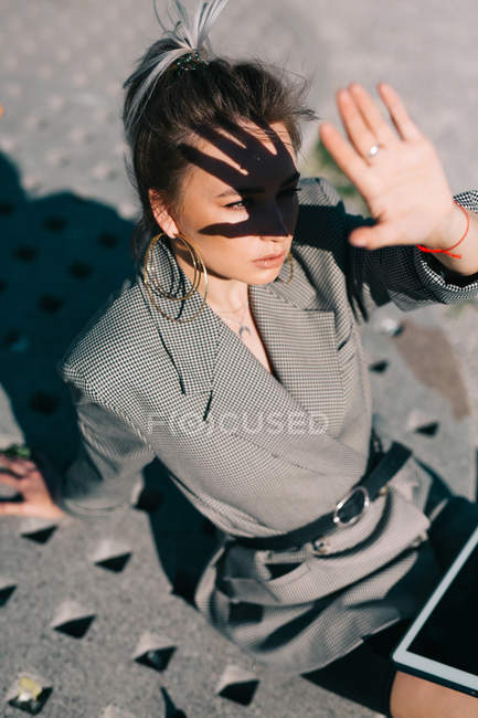 Стильна бізнес-леді, що працює на планшеті, сидячи на квадратній тарілці і піднімаючи руку, щоб закрити обличчя від яскравого сонця — стокове фото