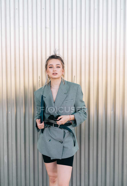 Trendy businesswoman holding tablet and sunglasses, leaning on metal wall and looking at camera with questioning expression — Stock Photo