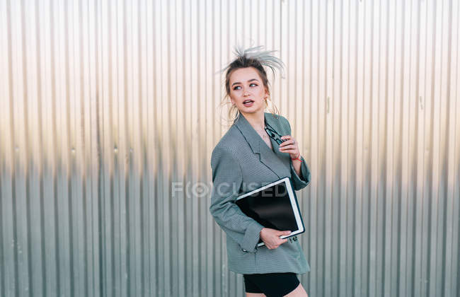 Trendy businesswoman holding tablet and sunglasses, leaning on metal wall and looking away with questioning expression — Stock Photo