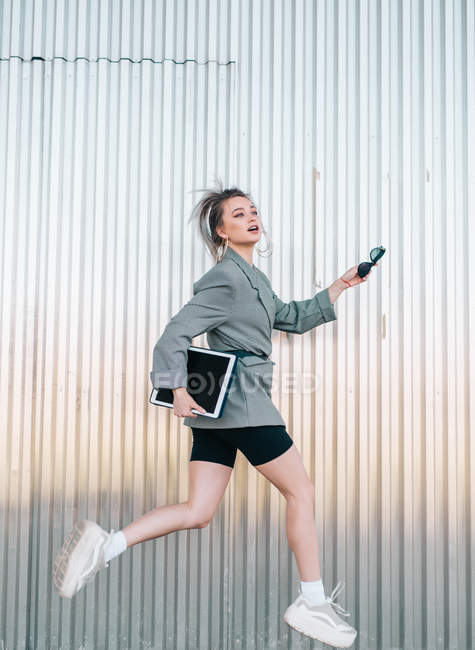 Mulher de negócios na moda segurando tablet e óculos de sol, pulando na parede de metal e olhando para longe — Fotografia de Stock