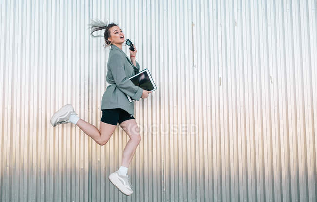 Mulher de negócios na moda segurando tablet e óculos de sol, pulando na parede de metal e olhando para longe — Fotografia de Stock