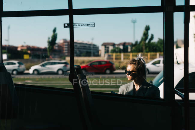 Femme d'affaires branchée avec des lunettes de soleil penché derrière le bus et regardant loin — Photo de stock