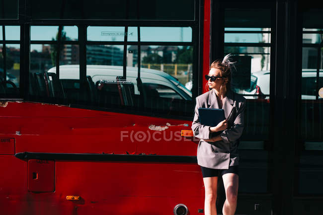 Donna d'affari alla moda che tiene tablet, indossa tuta e occhiali da sole, appoggiata sul bus rosso e distoglie lo sguardo — Foto stock