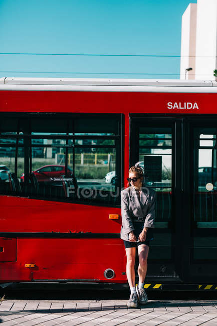 Trendige Geschäftsfrau hält Tablet in der Hand, trägt Anzug und Sonnenbrille, lehnt an rotem Bus und schaut weg — Stockfoto