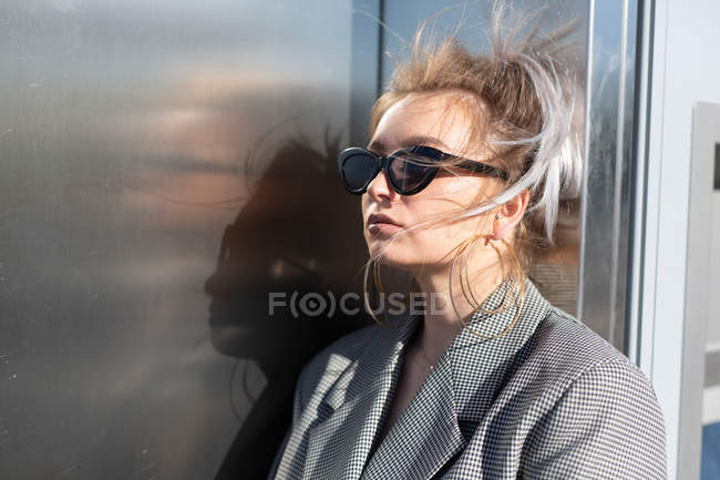 Donna attraente alla moda con acconciatura alla moda e occhiali da sole nelle vicinanze parete in metallo scuro lucido guardando altrove — Foto stock