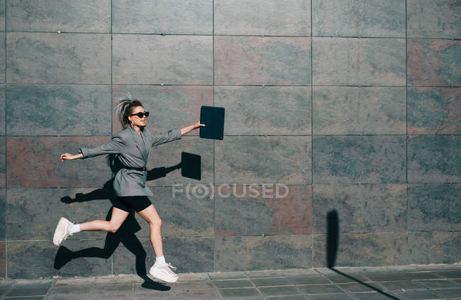 Seitenansicht einer stilvollen Geschäftsfrau, die entlang der Marmorwand springt und mit Tablet winkt — Stockfoto