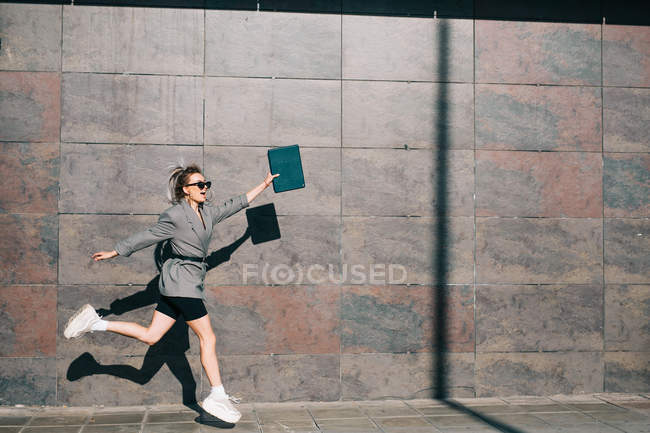 Vista laterale di elegante donna d'affari saltando lungo la parete di marmo e agitando le mani con tablet — Foto stock