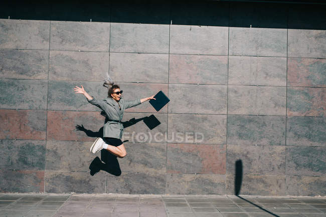 Side view of stylish businesswoman jumping along marble wall and waving hands with tablet — Stock Photo