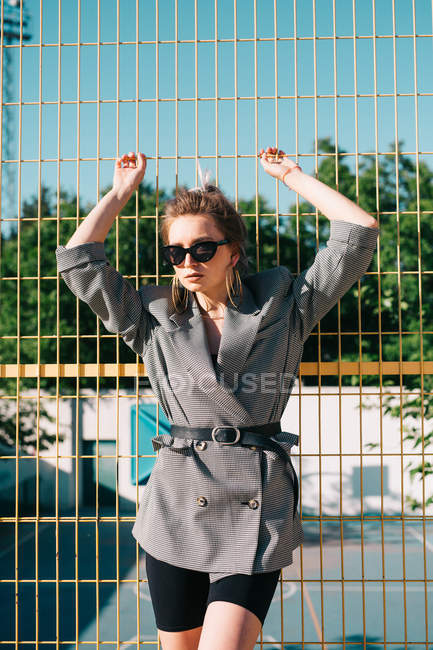 Femme d'affaires à la mode à la mode regardant la caméra dans les lunettes de soleil et s'appuyant sur un treillis jaune dans une journée lumineuse sur une aire de jeux — Photo de stock