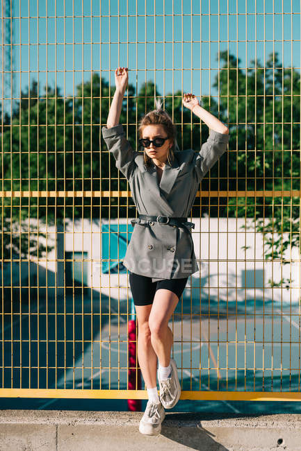 Trendy fashionable businesswoman looking at camera in sunglasses and leaning on yellow lattice in bright day on playground — Stock Photo