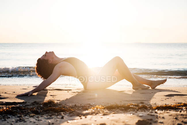 Босоногая женщина в купальниках, откидывающаяся назад и закрывающая глаза, сидя на мокром песке рядом с морем, освещенном сзади — стоковое фото