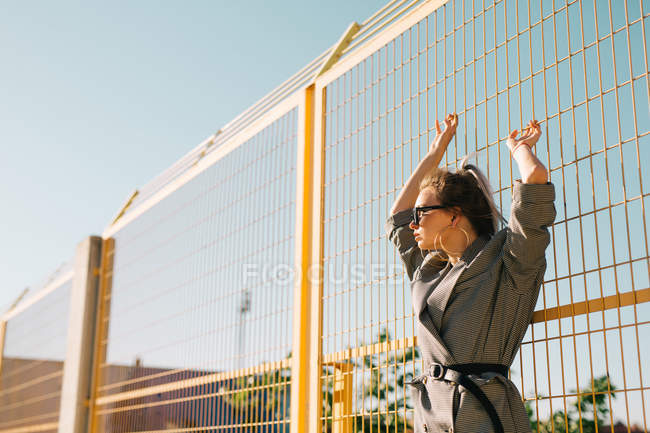 Tendance femme d'affaires à la mode regardant loin dans les lunettes de soleil et s'appuyant sur treillis jaune dans la journée brillante sur aire de jeux — Photo de stock
