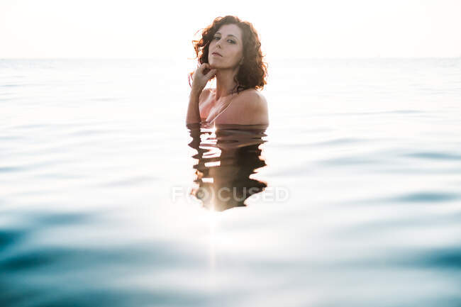 Vista lateral da bela senhora olhando para a câmera enquanto está em pé na água do mar limpa no dia ensolarado — Fotografia de Stock