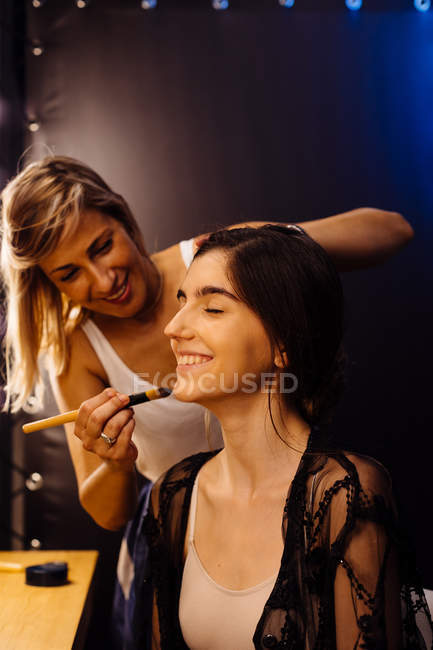 Vista lateral do estilista aplicando maquiagem no modelo morena sentado na frente do espelho iluminado no vestiário — Fotografia de Stock