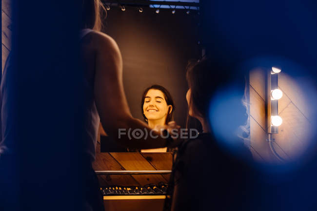 Vista trasera de estilista aplicando maquillaje a modelo morena sentada frente a espejo iluminado en vestidor - foto de stock