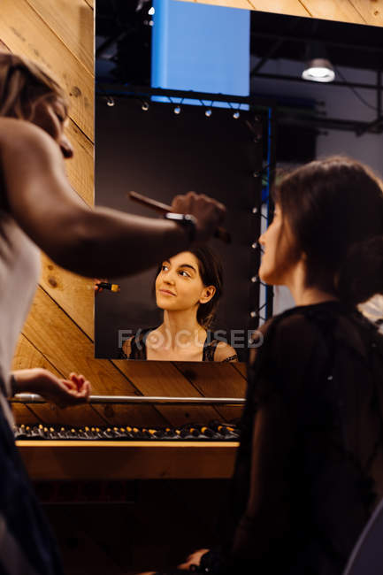 Vue arrière du styliste appliquant du maquillage sur le modèle brune assis devant un miroir éclairé dans le vestiaire — Photo de stock