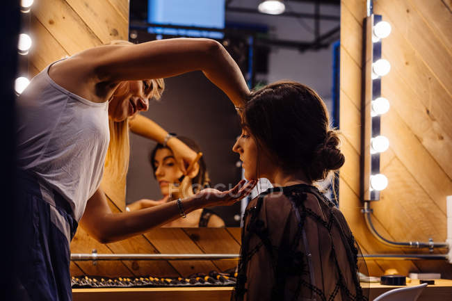 Visão traseira do estilista aplicando maquiagem no modelo morena sentado na frente do espelho iluminado no vestiário — Fotografia de Stock