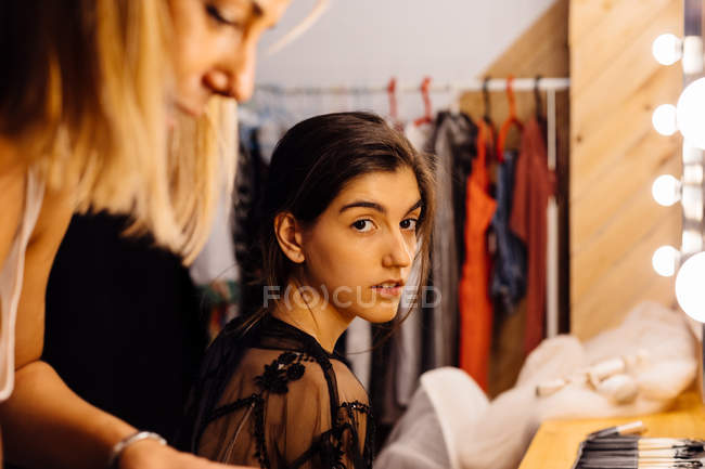 Vista laterale dello stilista che applica il trucco sul modello bruna seduto davanti allo specchio illuminato nello spogliatoio — Foto stock