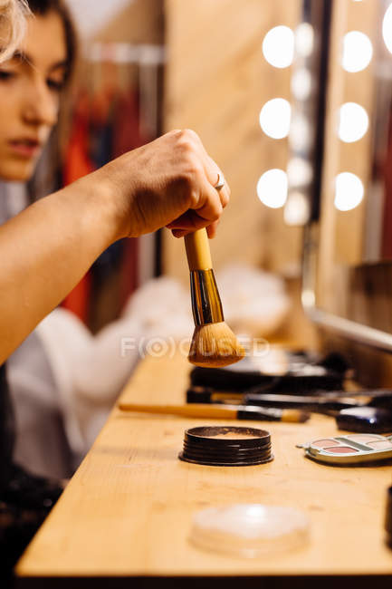 Vista lateral da mulher da colheita sentada na frente do espelho iluminado e escova de mascaramento estilista em pó para fazer maquiagem — Fotografia de Stock
