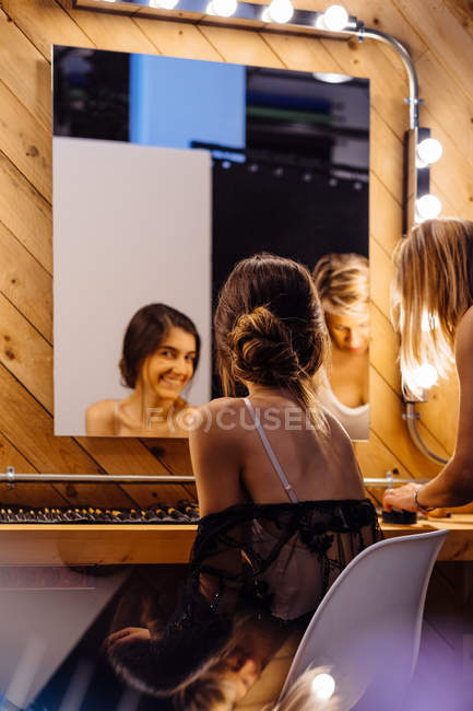 Visão traseira do estilista aplicando maquiagem no modelo morena sentado na frente do espelho iluminado no vestiário — Fotografia de Stock