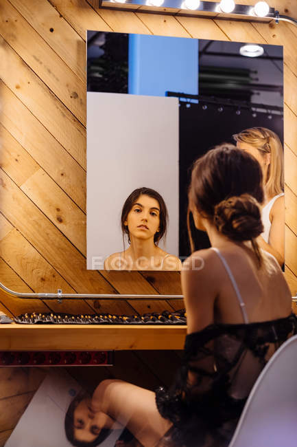 Rückansicht einer glücklichen jungen Frau, die sich im Spiegel spiegelt, der an der Holzwand hängt, während sie in der Garderobe sitzt — Stockfoto