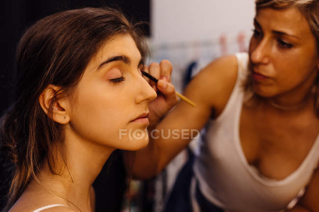 Seitenansicht einer brünetten Frau, die mit geschlossenen Augen sitzt, während ein Visagist auf verschwommenem Hintergrund Make-up aufträgt — Stockfoto