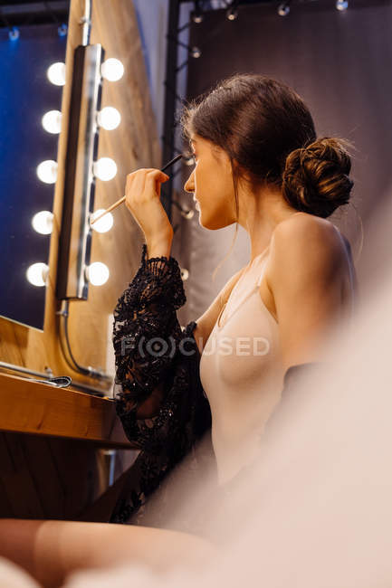 Side view of young glamorous woman applying eye shadows while sitting by illuminated mirror and doing makeup in dressing room — Stock Photo