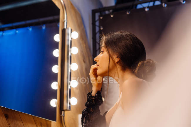 Вид сбоку на привлекательную женщину с темными волосами в черном прозрачном платье, делающую макияж, сидя перед освещенным зеркалом — стоковое фото