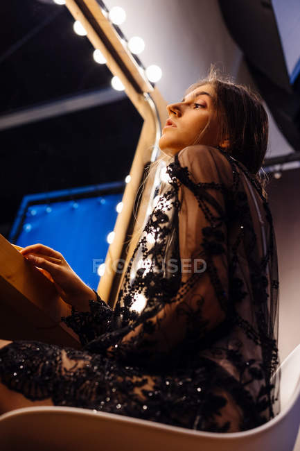 Dal basso vista laterale di attraente donna bruna in pizzo vestito nero seduto sulla sedia guardando allo specchio e preparandosi — Foto stock