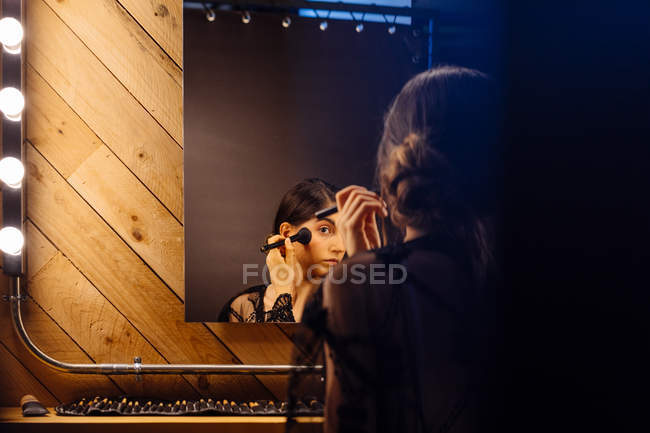 Задний вид привлекательной женщины с темными волосами в черном прозрачном платье делает макияж, сидя перед освещенным зеркалом — стоковое фото