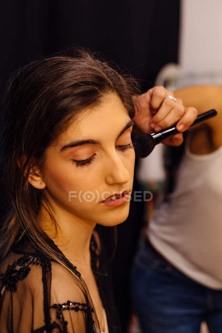 Vista lateral do estilista aplicando maquiagem no modelo morena sentado na frente do espelho iluminado no vestiário — Fotografia de Stock