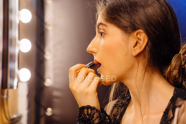 Side view of serious brunette woman in lace black dress applying red lipstick while doing makeup in dressing room — Stock Photo