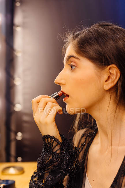 Seitenansicht einer ernsten brünetten Frau in einem schwarzen Spitzenkleid, die roten Lippenstift aufträgt, während sie in der Garderobe Make-up macht — Stockfoto