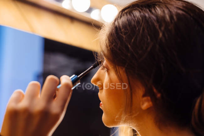 Vista lateral de la cosecha morena atractiva mujer mirando en el espejo haciendo maquillaje aplicando rímel en las pestañas - foto de stock