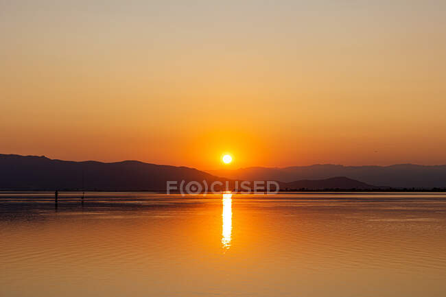 Orange sun going down behind dark hills reflecting in peaceful slightly rippled water creating romantic seascape — Stock Photo