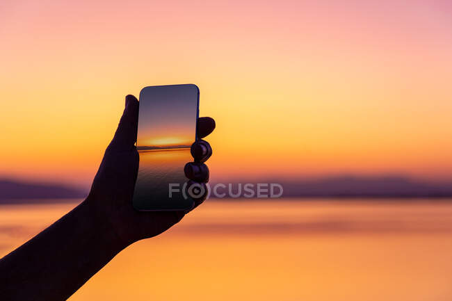 Faceless tourist holding smartphone with shot of beautiful orange sunset on blurred background — Stock Photo