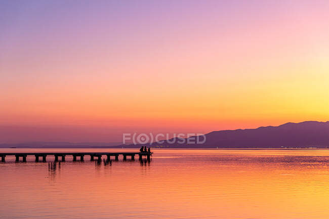 Silhouette di persone lontane sedute sul molo vicino al tranquillo lago contro il cielo senza nuvole del tramonto — Foto stock