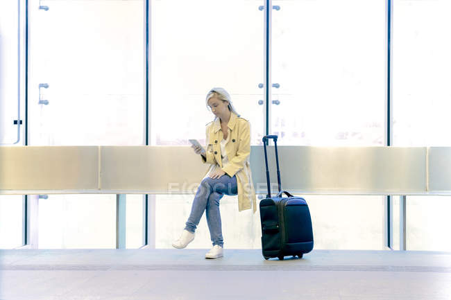 Юная блондинка-туристка пользуется смартфоном, сидя в аэропорту с рюкзаком — стоковое фото