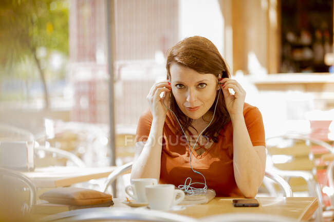 Erwachsene lächelnde Dame in Freizeitkleidung setzt Kopfhörer auf, während sie auf der gemütlichen Sommerterrasse des Cafés sitzt und sich ausruht — Stockfoto