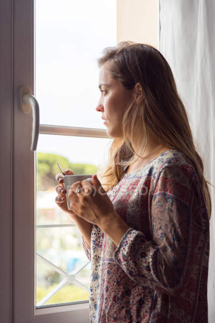 Nachdenkliches blondes Mädchen im Hemd mit einer Tasse Kaffee, das morgens aus dem Fenster schaut — Stockfoto
