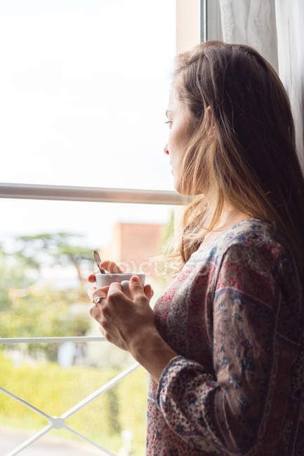 Menina loira pensativo na camisa com uma xícara de café olhando para as janelas da manhã — Fotografia de Stock