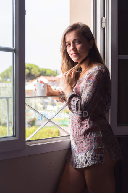 Menina loira pensativo na camisa com uma xícara de café olhando para a câmera em janelas da manhã — Fotografia de Stock