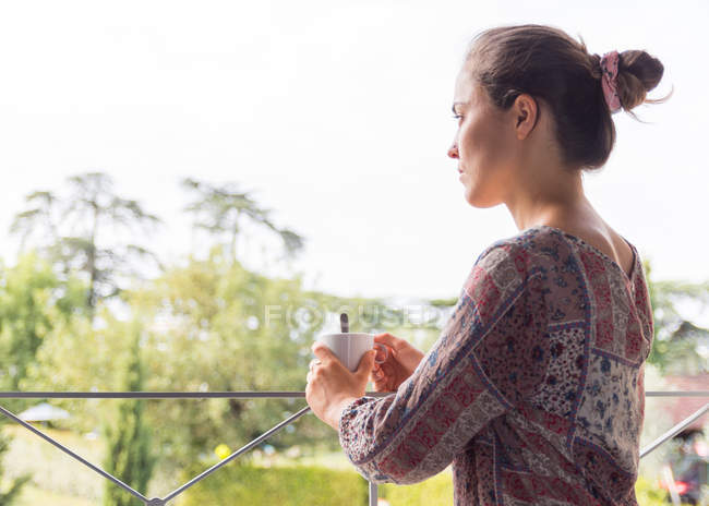 Красивая блондинка в рубашке с чашкой кофе, выглядывающая утром из окон — стоковое фото
