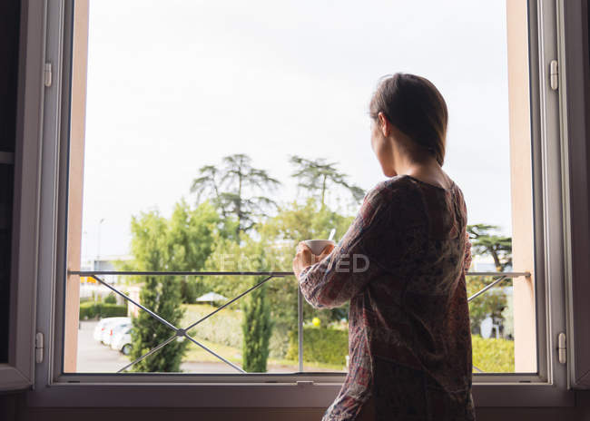 Menina loira pensativo na camisa com uma xícara de café olhando para as janelas da manhã — Fotografia de Stock